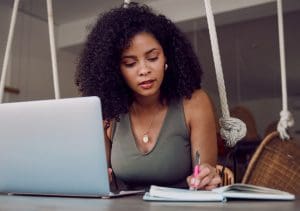 black woman in green shirt writing in notebook and silver laptop on desk
