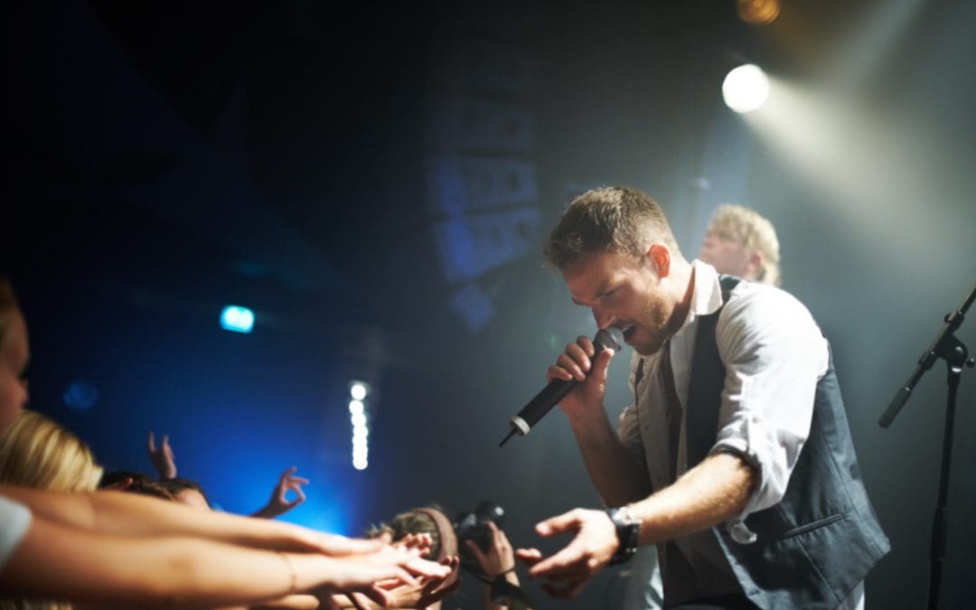 man singing on stage with mic in gray long sleeve and vest reaching out to fans in crowd